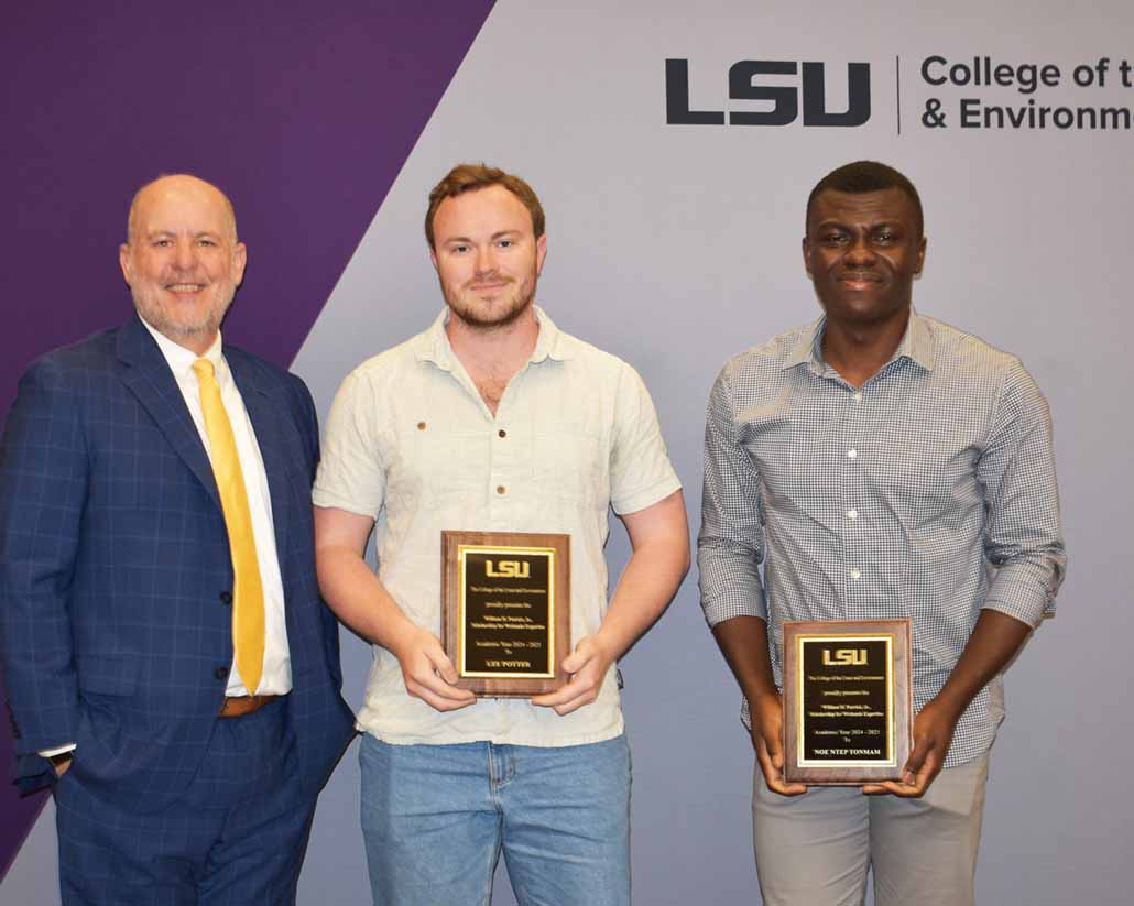 Two students dressed casually stand holding plaques next to a man in a suit